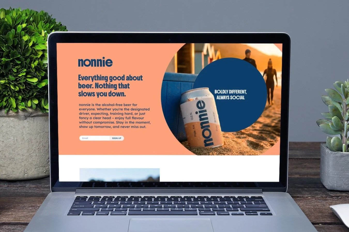 Laptop screen displays a webpage about nonnie, an alcohol-free beer, with a background image of people walking on a beach at sunset and a can of nonnie beer in the foreground. Potted plants sit on either side of the laptop.