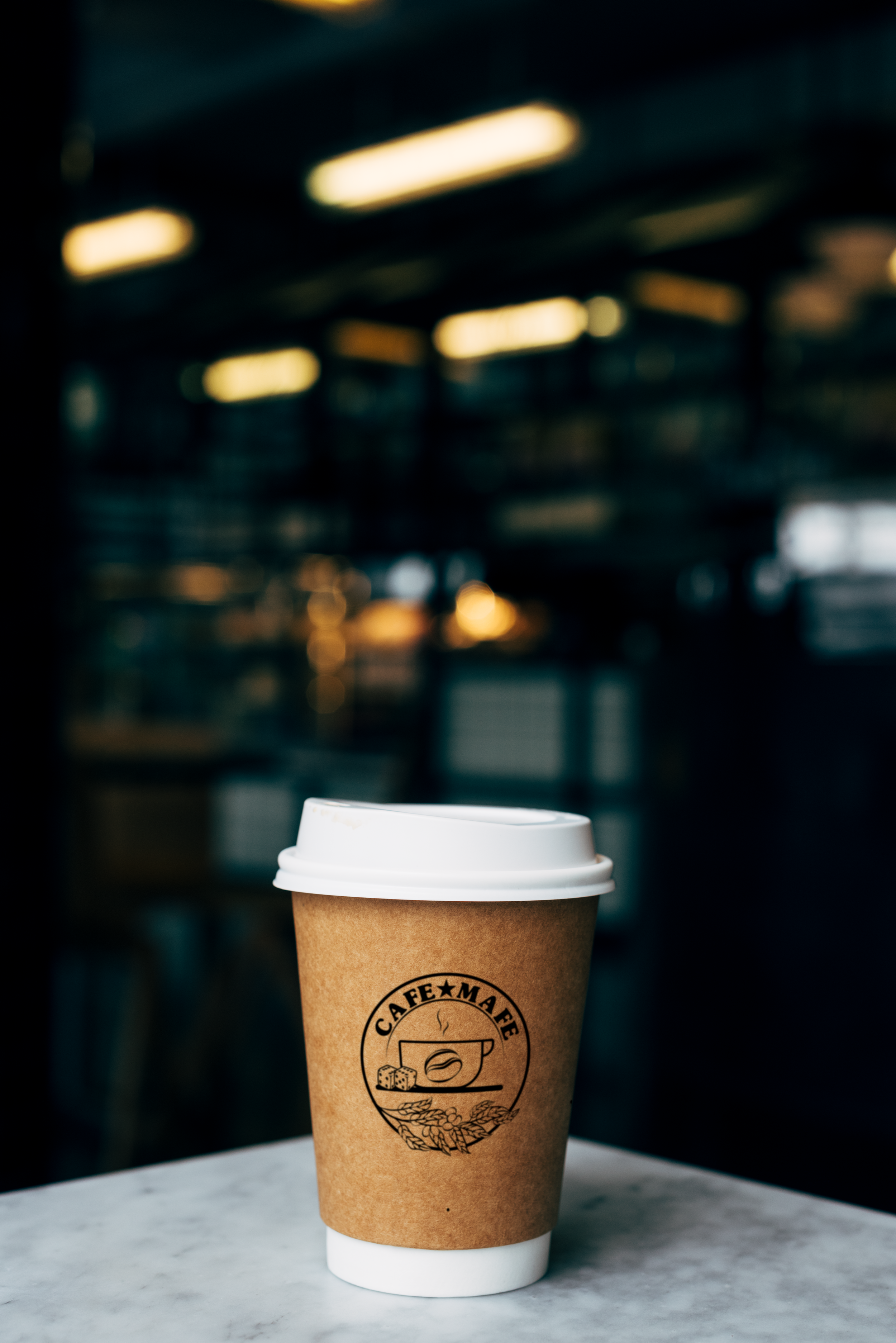 A disposable coffee cup with a white plastic lid and a brown cardboard sleeve sitting on a gray marble surface, with a blurred background of an industrial-style interior with overhead lights.