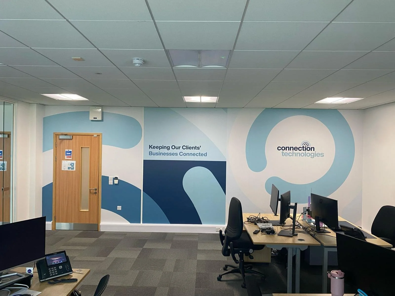 Office workspace with desks, computers, and chairs. A wall mural displays the company logo 'connection technologies' and the slogan 'Keeping Our Clients’ Businesses Connected.'