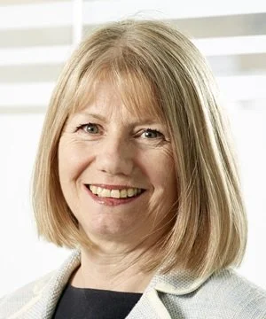 A woman with blonde hair smiling, wearing a light-colored blazer and black top, in front of a window with blinds.