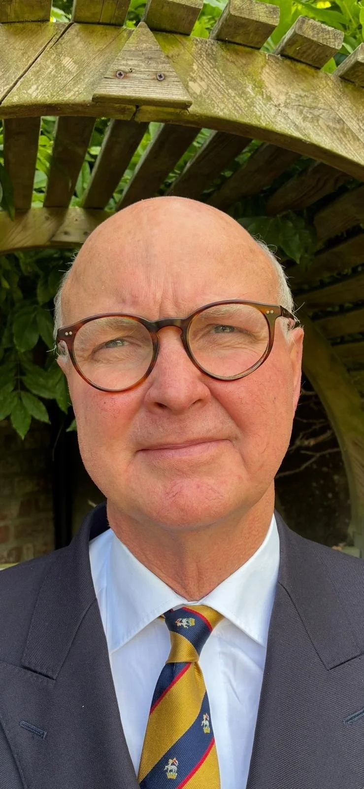 A man wearing a suit and tie taking a selfie outdoors, with a wooden structure and green foliage in the background.