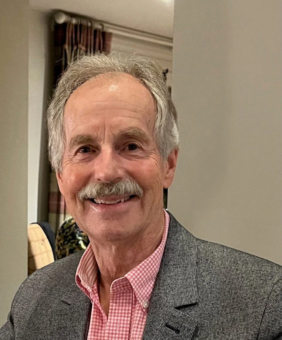 A smiling elderly man with gray hair and a mustache, wearing a gray blazer and pink checkered shirt, in an indoor setting with a window and curtains in the background.