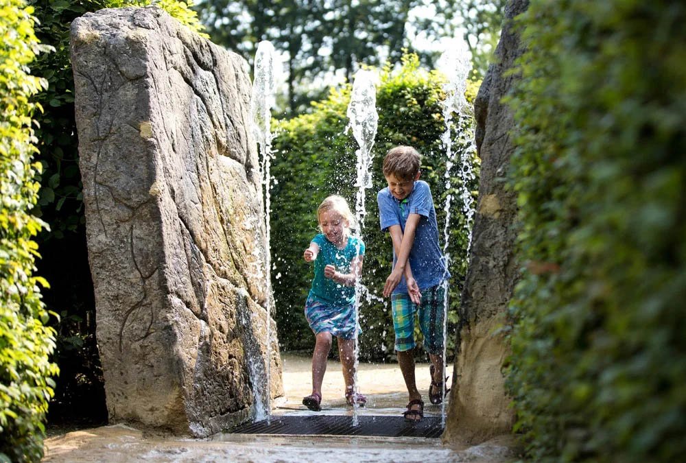 Twee kinderen spelen in een waterspel onder een fontein met grote stenen en groene struiken op de achtergrond.