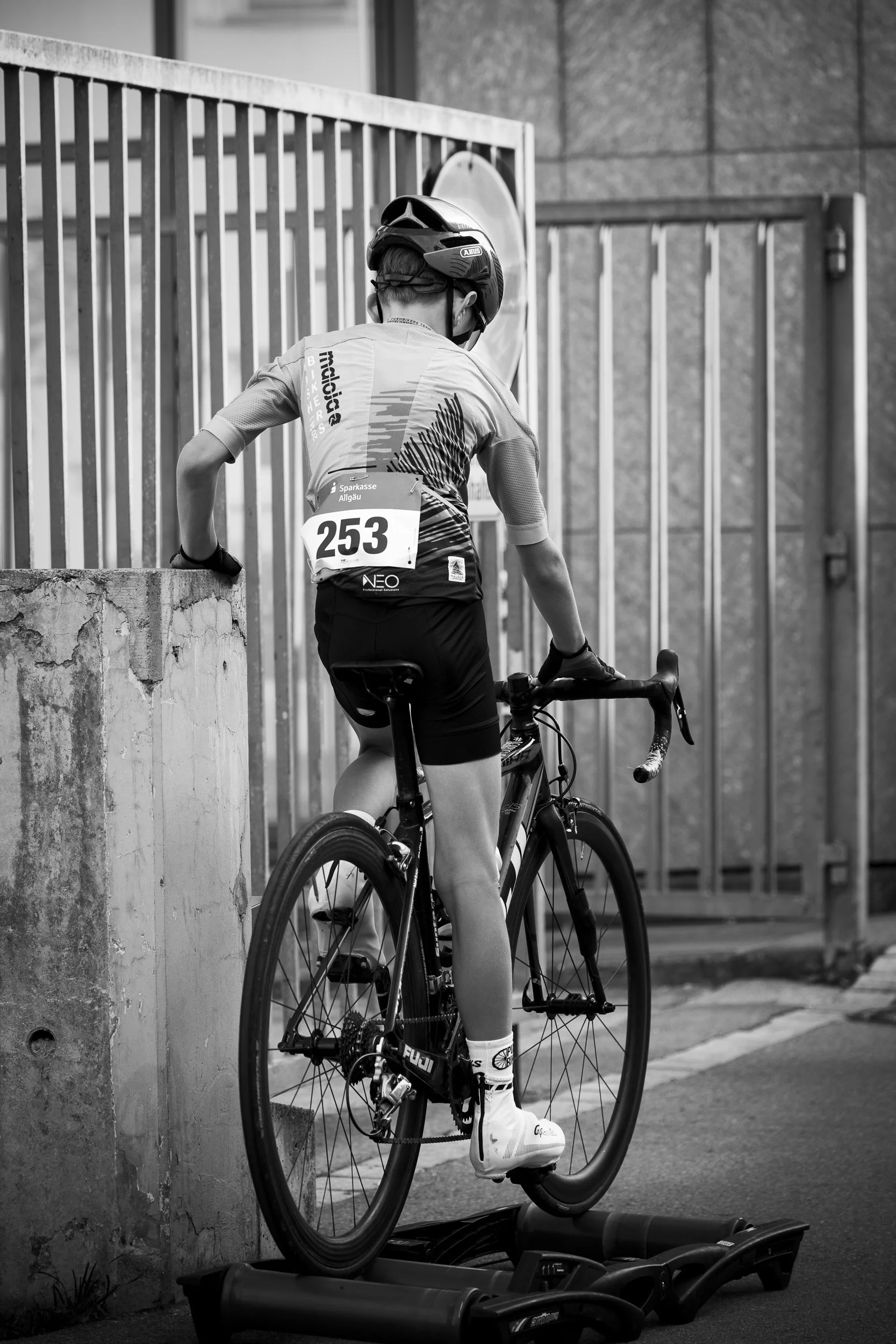 Ein junger Radfahrer mit Helm und Radnummer 253 steht mit seinem Fahrrad auf einem Rollentrainer, bereit für einen Wettkampf, in schwarz-weiß.