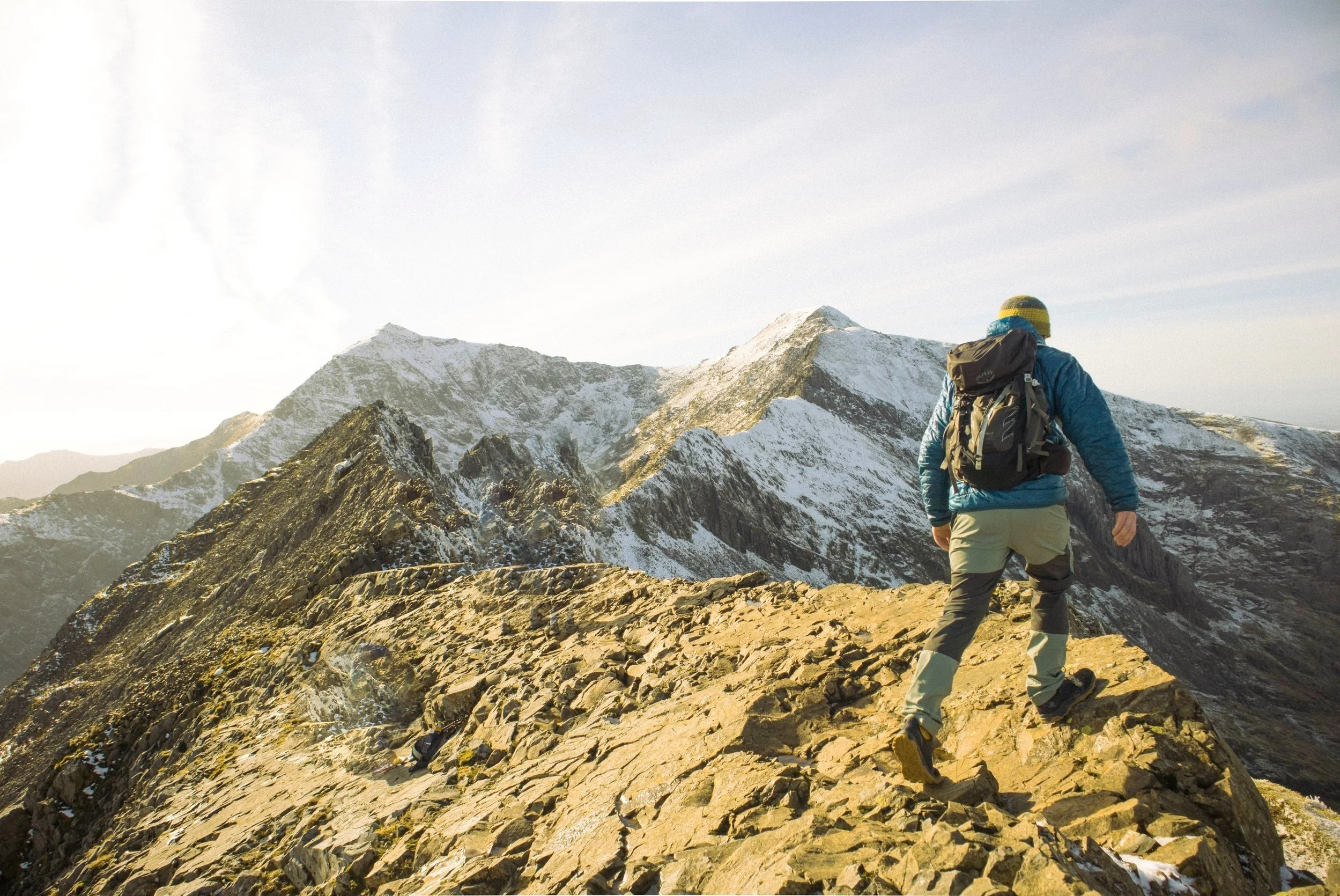 A hiker in outdoor gear walking on a rocky mountain ridge with snow-covered peaks in the background.