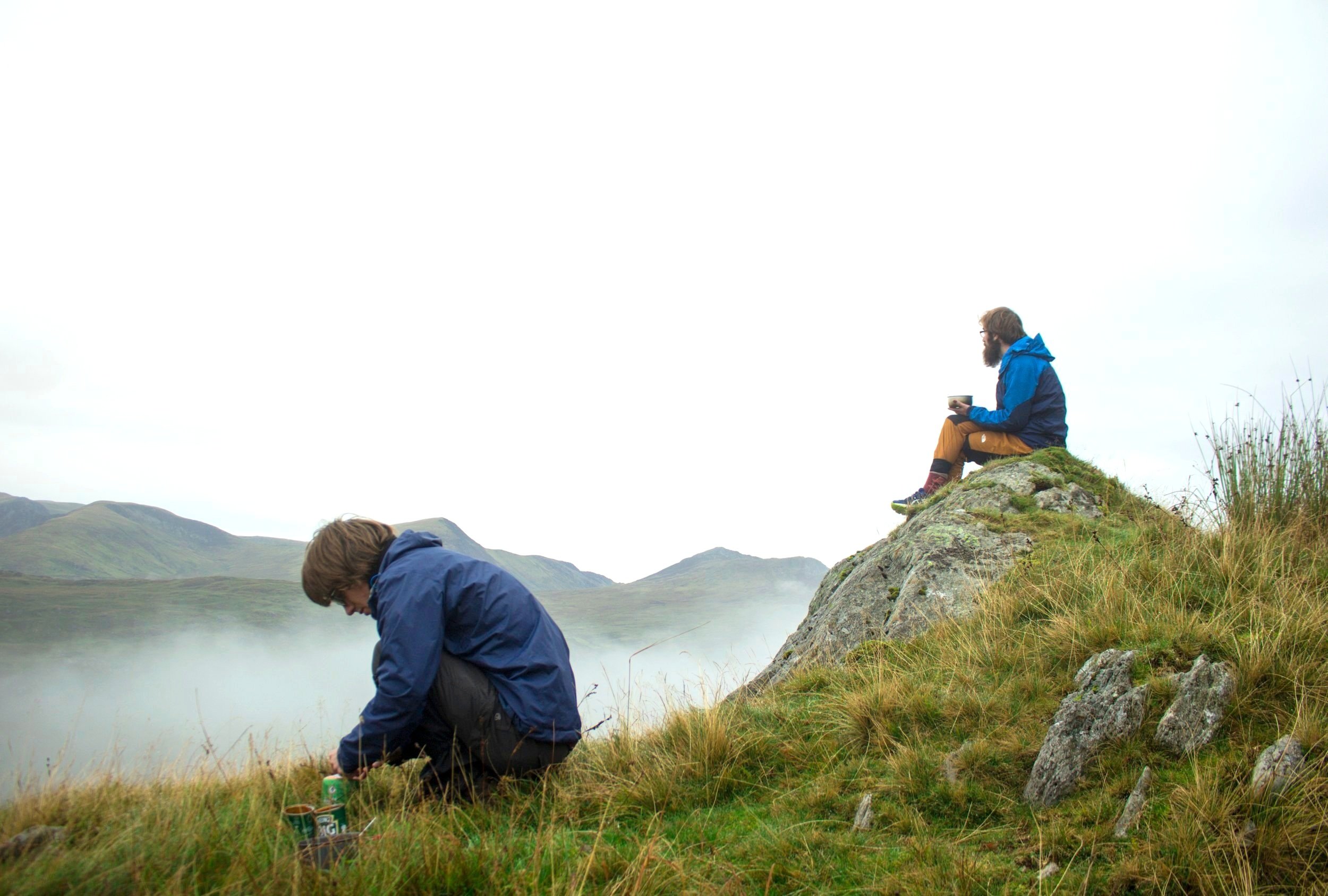 Two hikers in outdoor gear on a grassy hillside with mountains and fog in the background. One person is sitting on a rock eating and the other is crouched down, preparing breakfast.