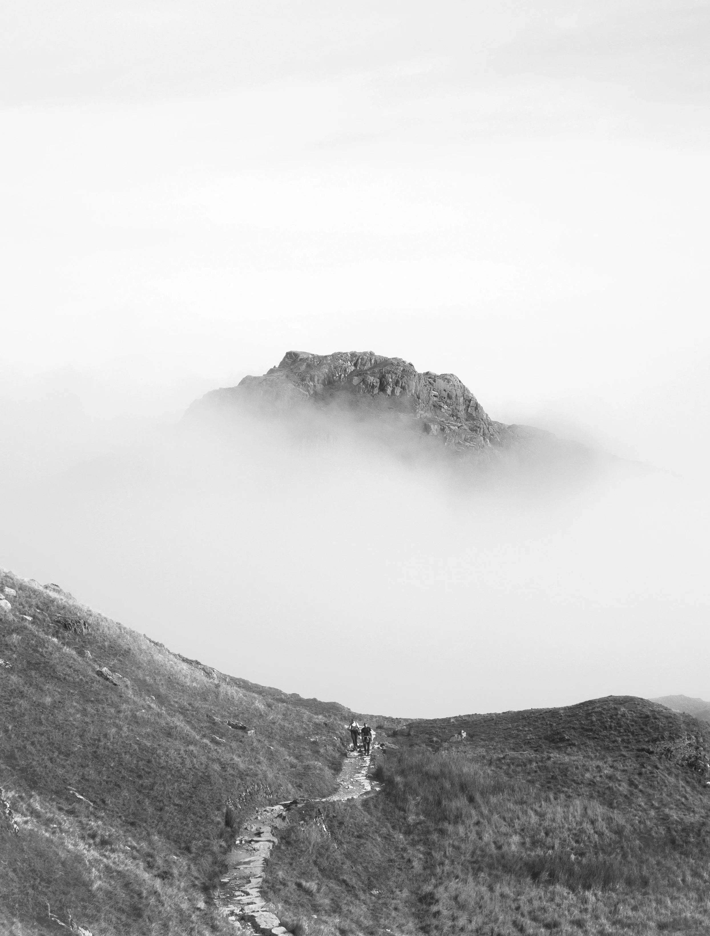 A black and white photo of a mountain landscape with hikers walking along a trail, fog partially obscuring the mountain.