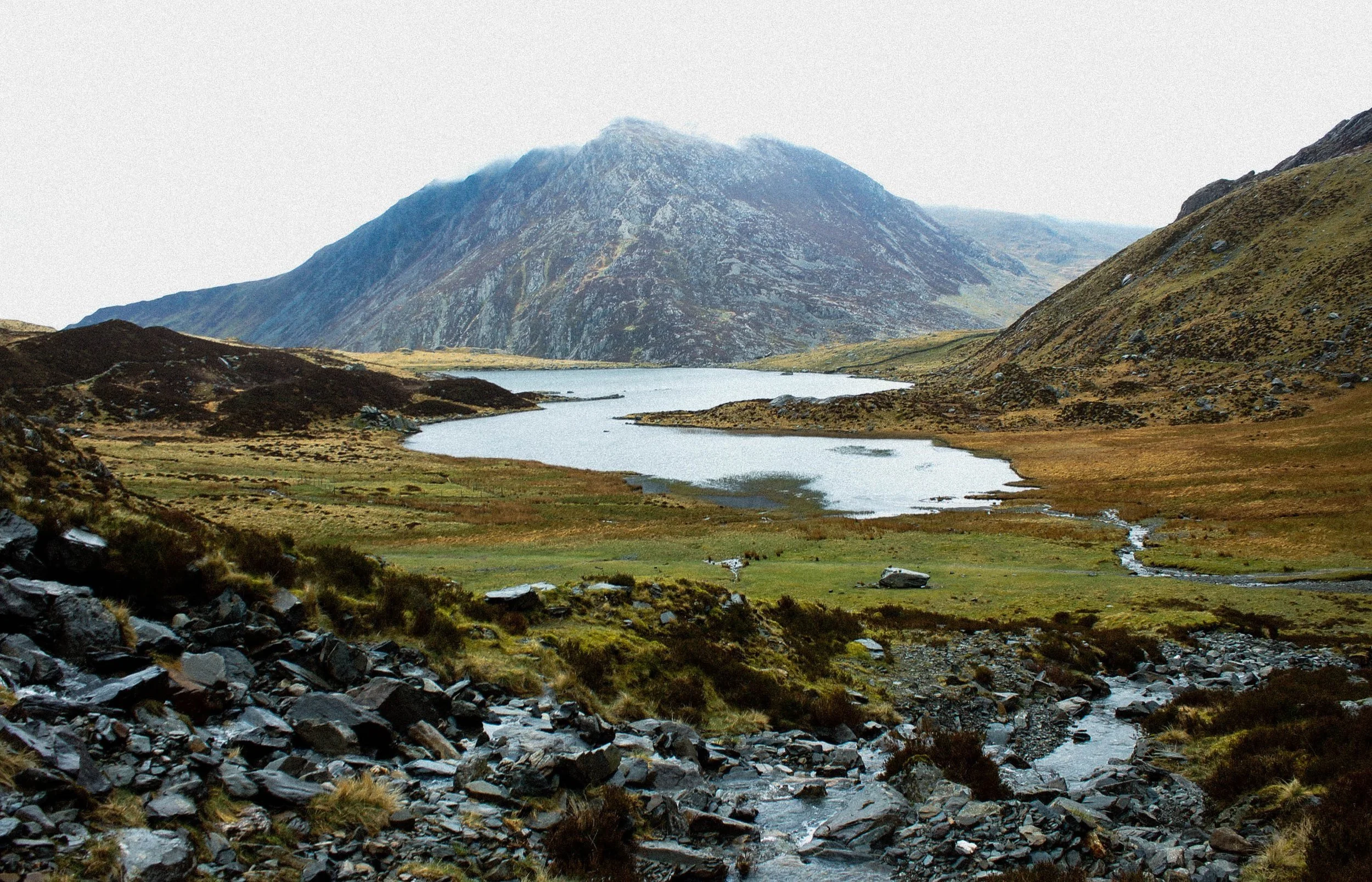 A mountain landscape featuring a lake with surrounding grassy and rocky terrain and distant mountains partially covered in clouds.