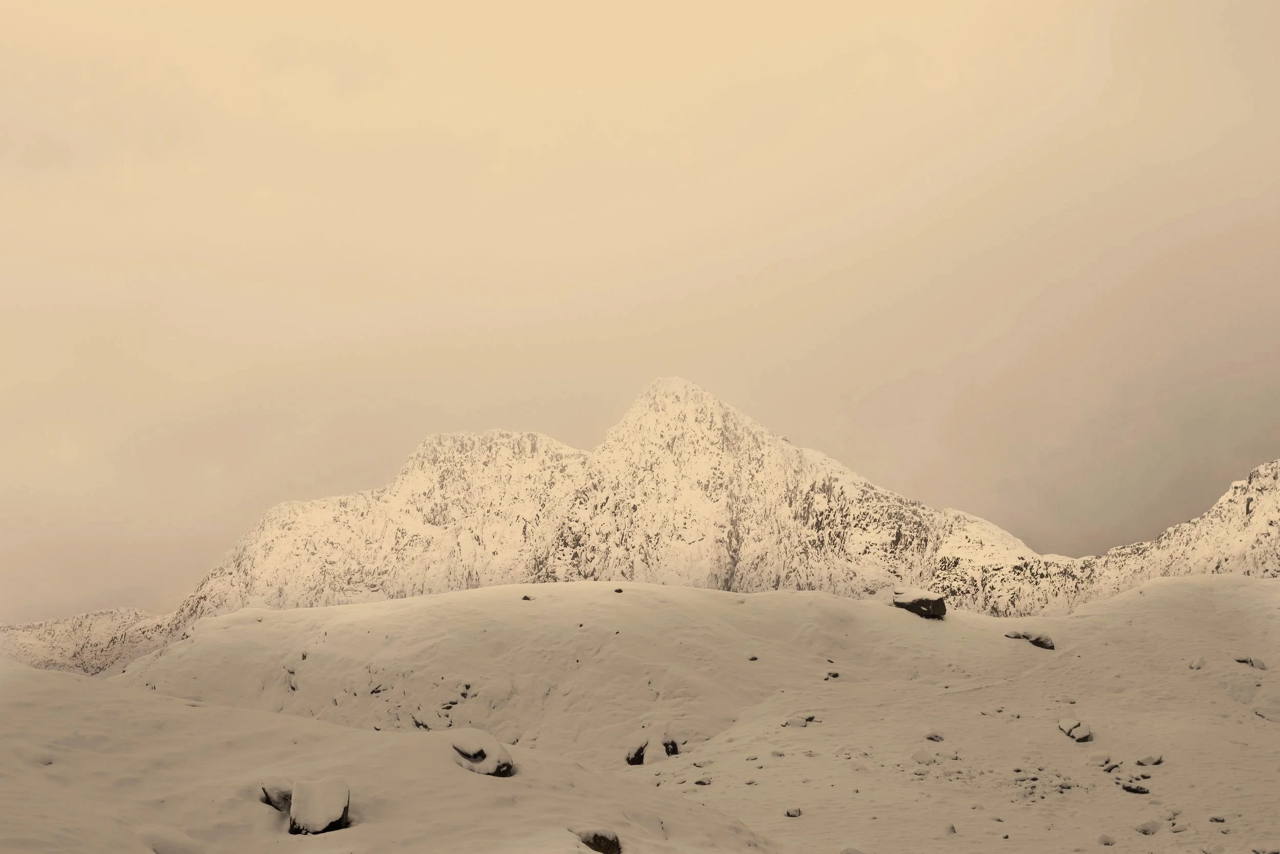 Snow-covered mountain range with a prominent peak in the background and a snow-blanketed landscape with rocks in the foreground.
