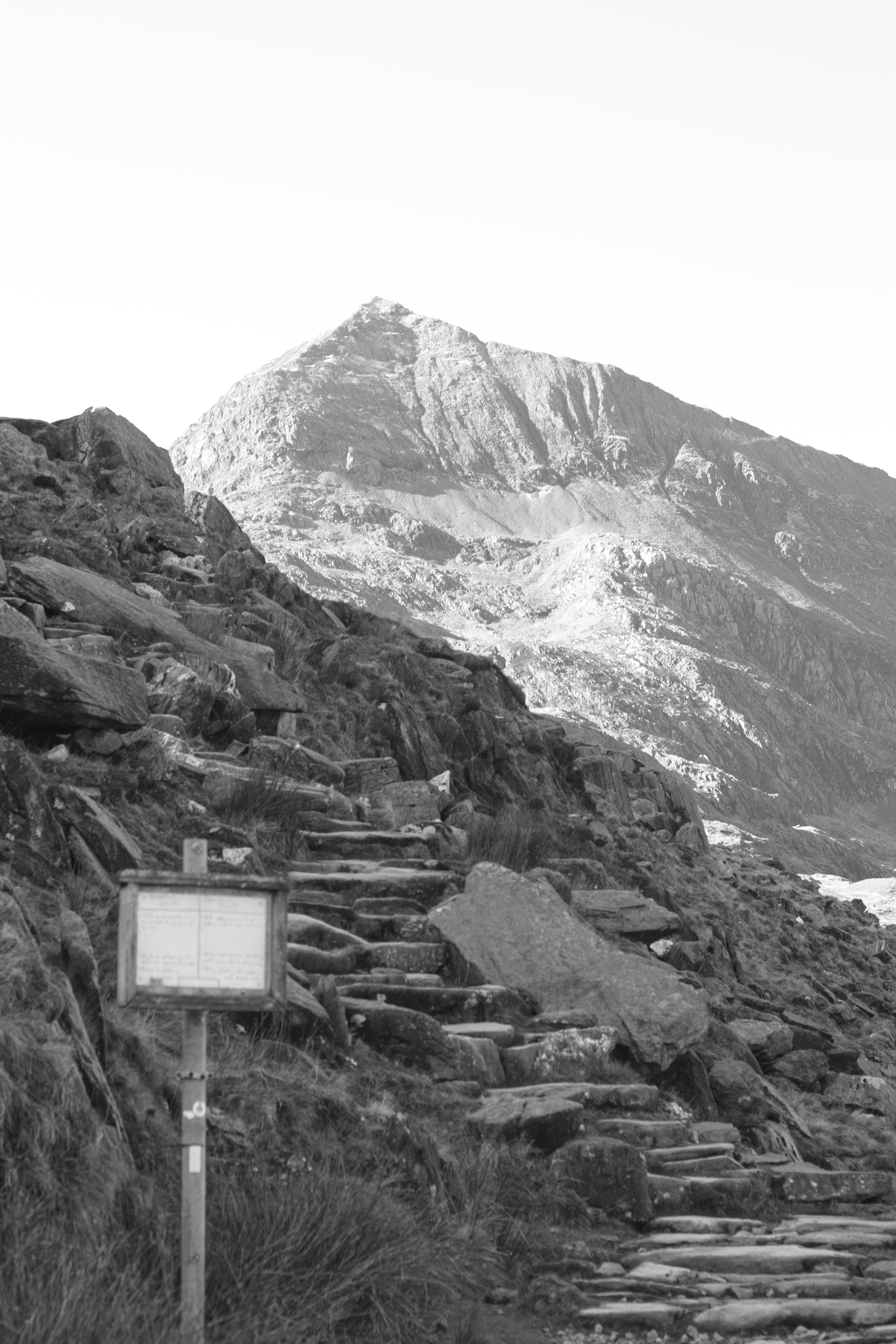A black-and-white photo of a mountain landscape with a rocky trail and steps leading up toward a prominent mountain peak.