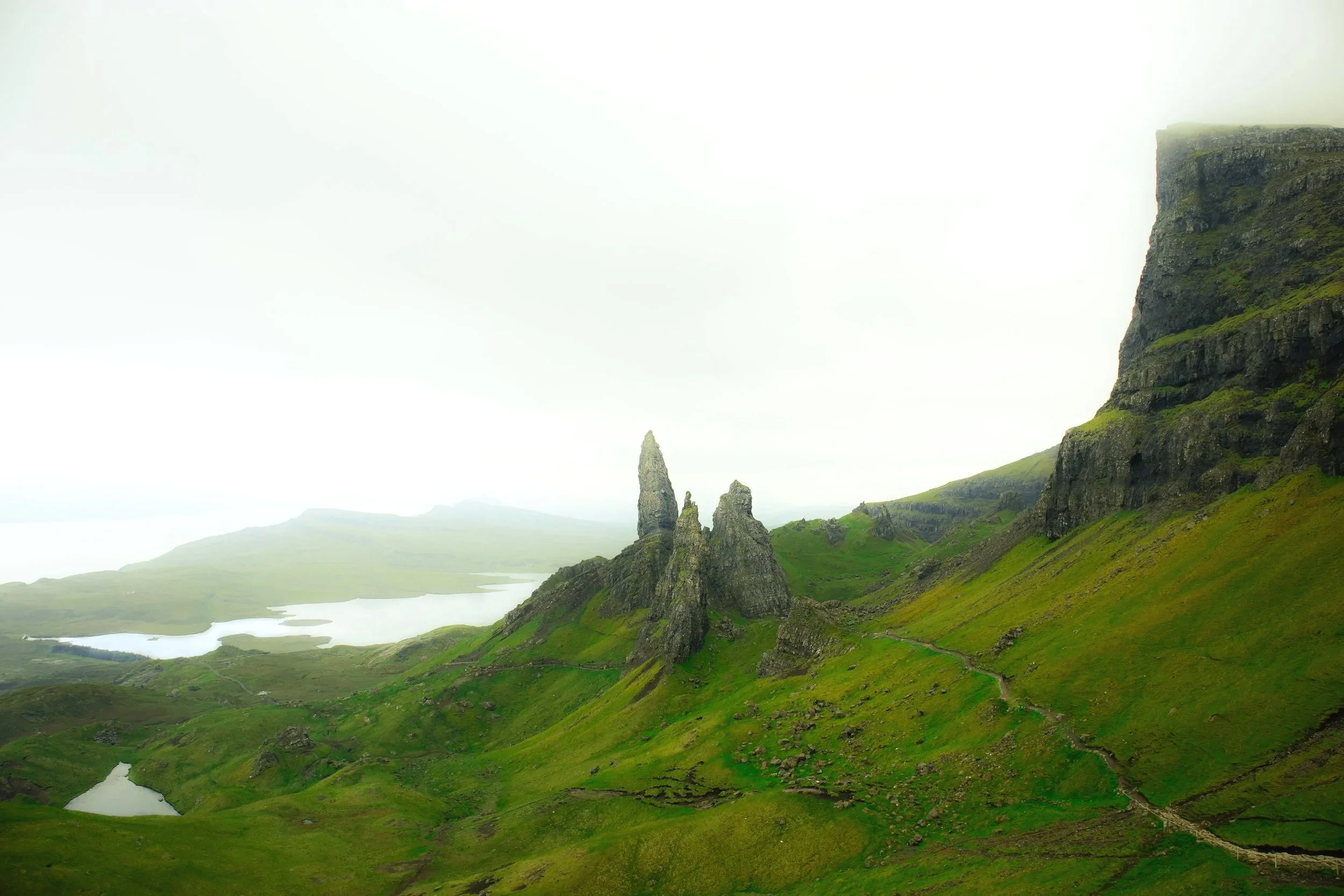Green landscape with rocky formations and cliffs, foggy sky, and small lakes in the distance.
