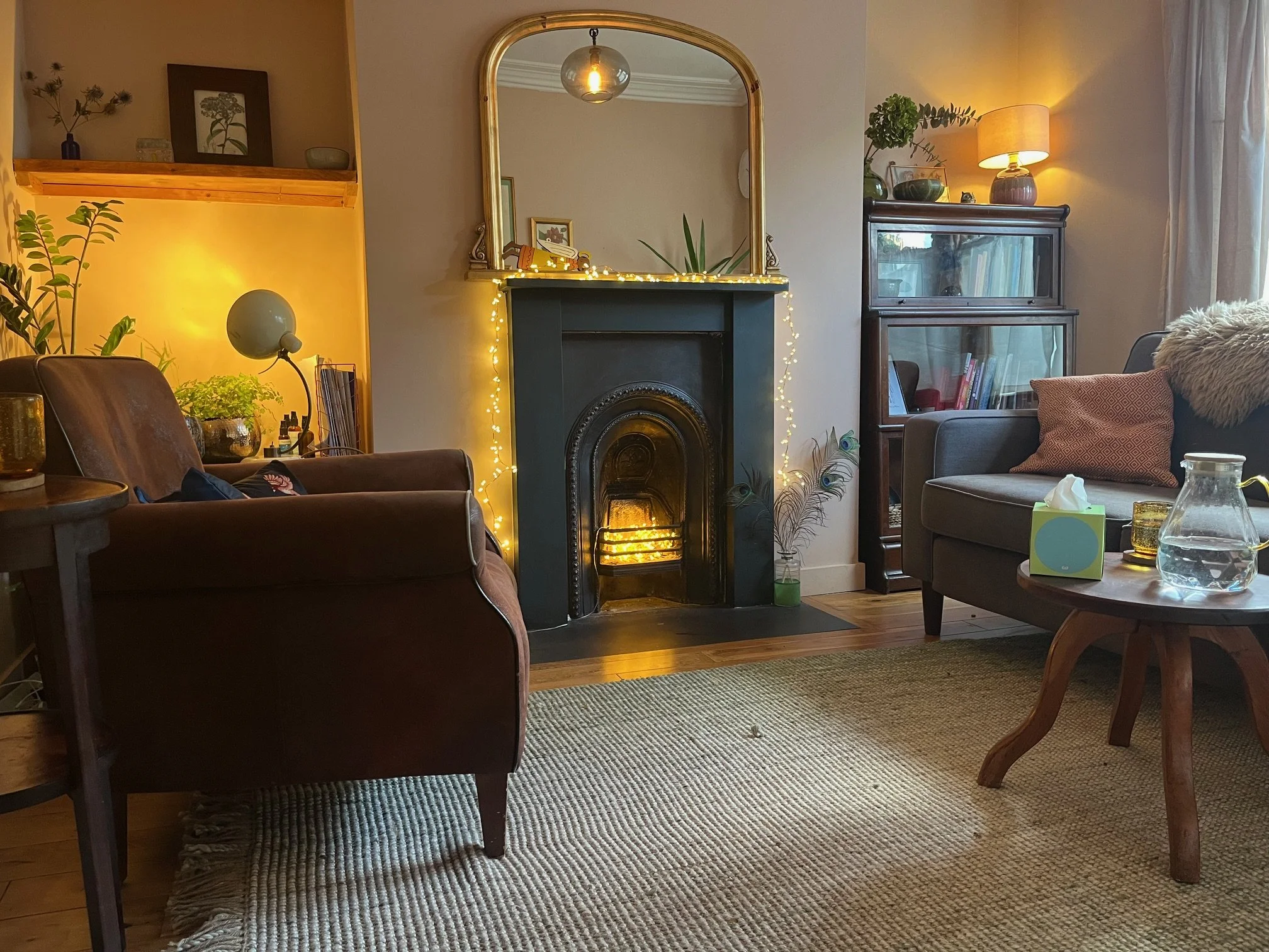 A cozy living room with a fireplace decorated with string lights, a large mirror above, and a burning fire inside. There are two couches, a coffee table with tissues and a glass pitcher, and various plants and decorations around the room.