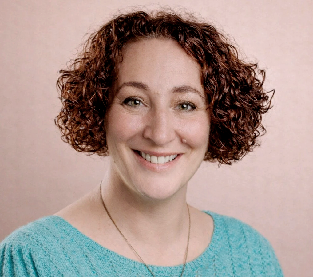Portrait of a woman with short, curly red hair, wearing a turquoise sweater and a necklace, smiling at the camera against a light pink background.
