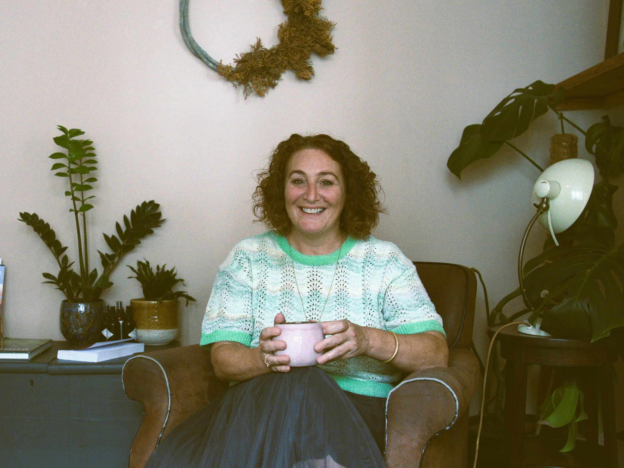 A woman with curly red hair smiling and holding a pink mug while sitting on a brown armchair in a room with green plants and a white wall with a decorative circular wreath.