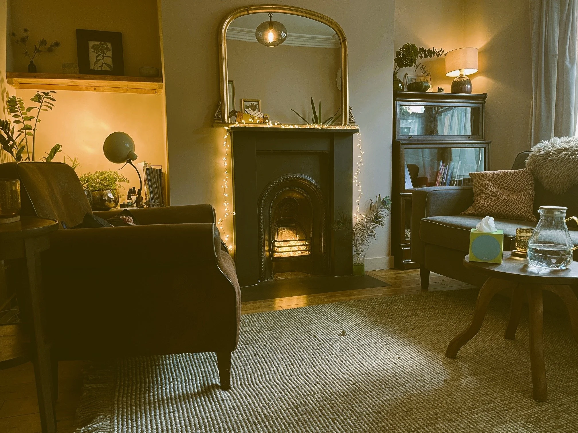 Cozy living room with a lit fireplace, mirror above, and string lights, featuring armchairs, a round coffee table with tissue box and pitcher, bookshelf with books, and various decorative plants and lamps.