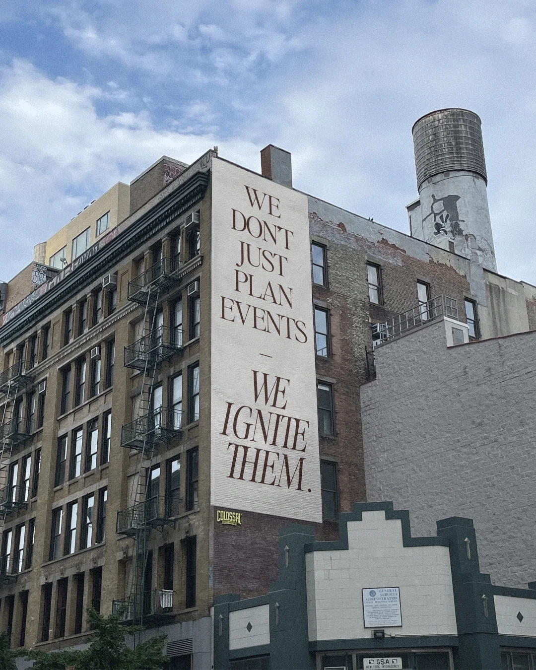 Large vertical sign on building wall with the message: 'We don't just plan events - we ignite them.' The building is located in an urban area with fire escapes on its side, and there is a chimney and graffiti visible on nearby structures.