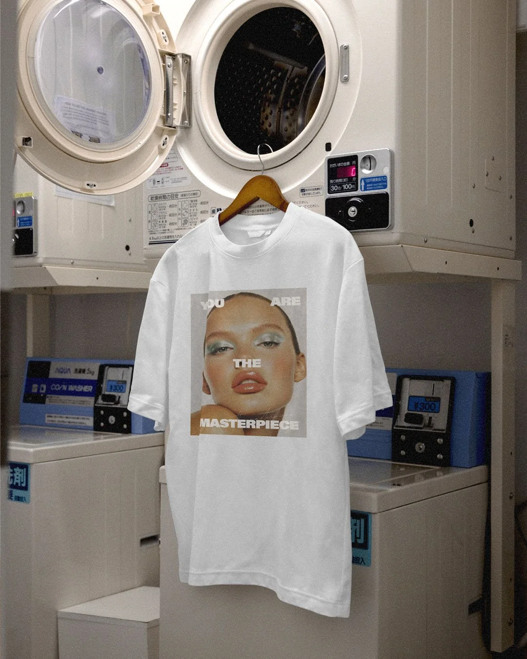 A white T-shirt with a printed image of a woman's face hanging on a wooden hanger in front of a laundry dryer's open door in a laundromat.