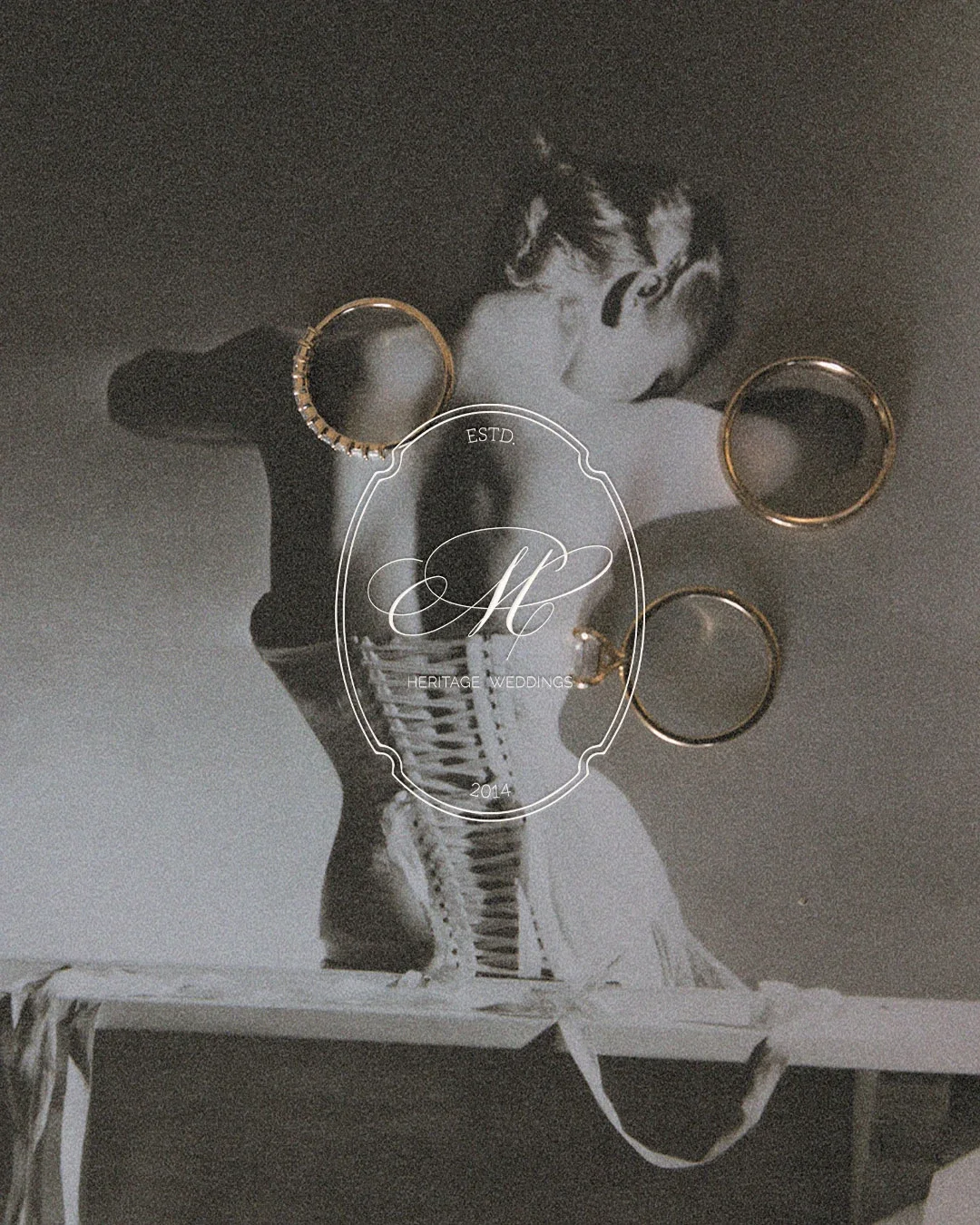 Black and white photograph of the back of woman in a corset, with three gold rings and a logo-overlay reading 'Heritage Weddings ESTD 2014'.