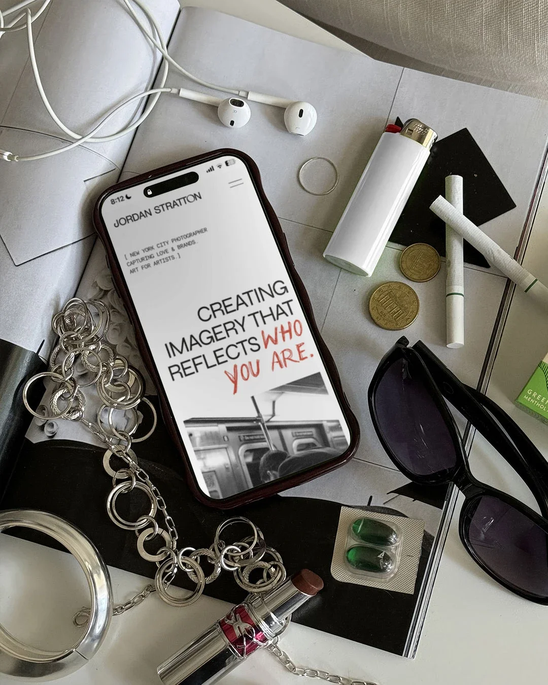 A smartphone showing a quote that reads "creating imagery that reflects who you are" surrounded by various personal items including sunglasses, coins, pills, a lighter, jewelry, a pen, an earphone, and a packet of gum, all on a light-colored surface.