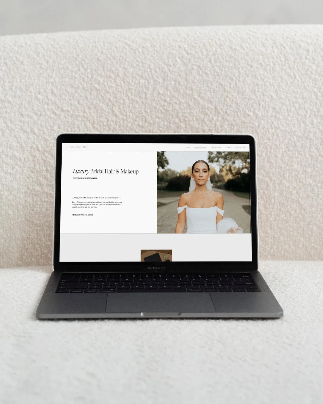 Open MacBook Pro on a white textured surface displaying a webpage for luxury bridal hair and makeup with a photo of a bride in a white dress and veil outdoors.