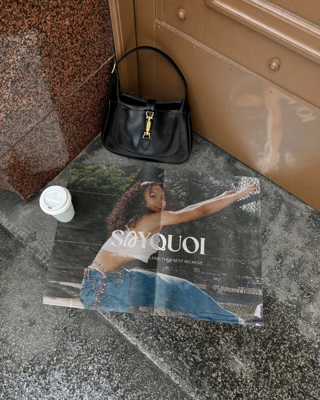 A black handbag, a disposable coffee cup, and a fashion magazine with a woman in denim shorts and a white top are on a sidewalk against a brick wall and a brown door.