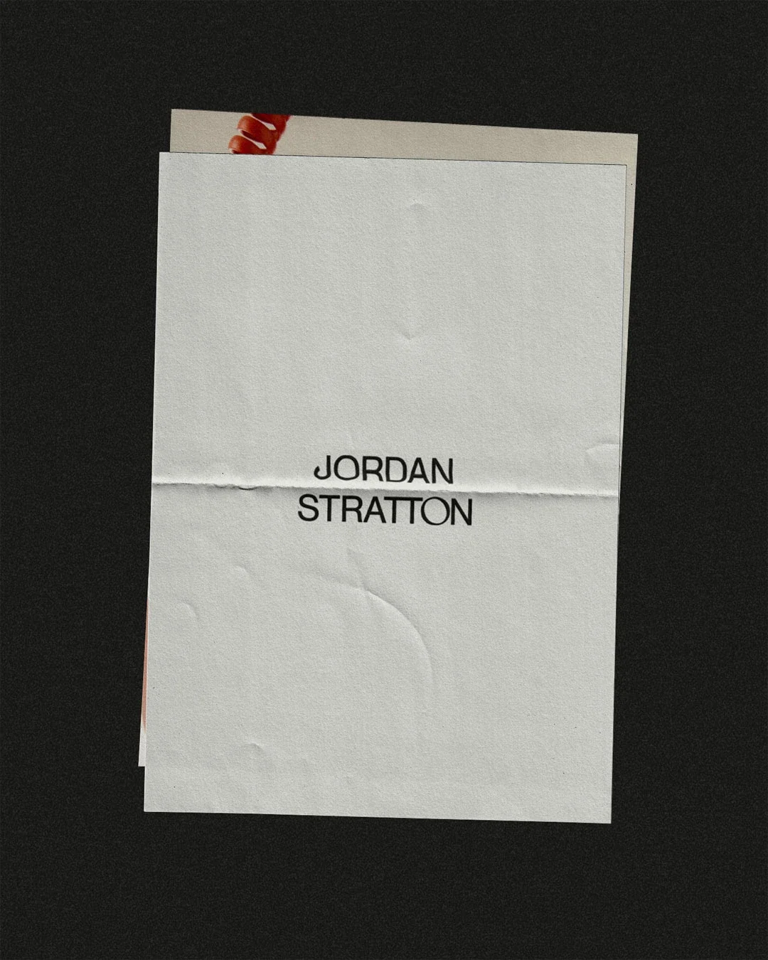 A piece of white paper with the name 'JORDAN STRATTON' written in black, placed on a dark surface. Part of another paper is visible underneath.