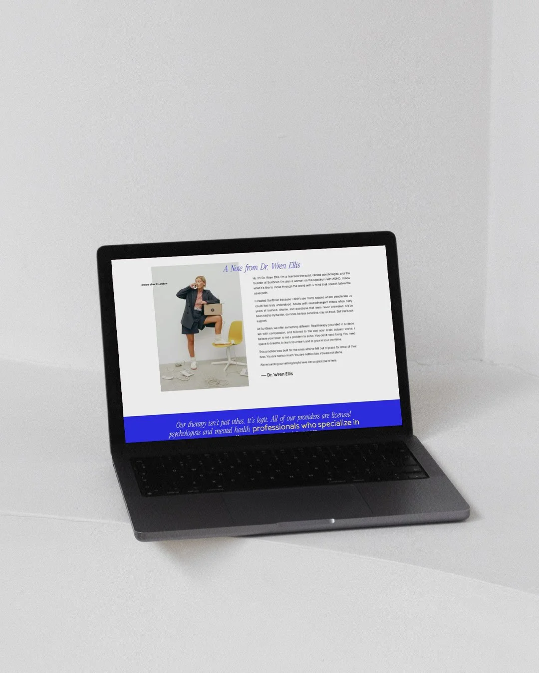 A black laptop on a white table displaying a webpage titled "A Note from Dr. Wren Ellis" with an image of a woman in a blazer and shorts, sitting on a yellow chair with a laptop and a coffee mug, and text about mental health and psychology.