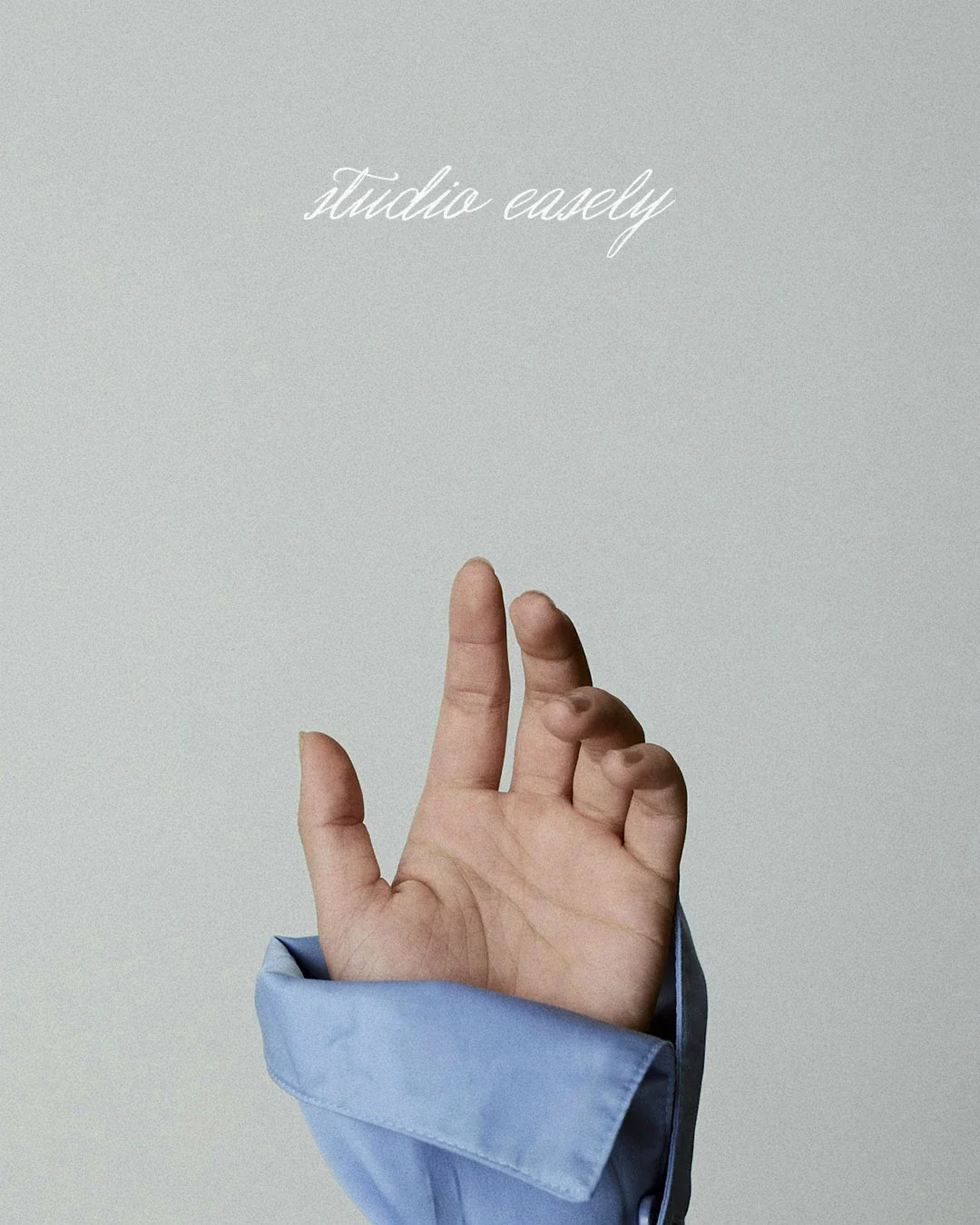 Close-up of a hand with fingers in a delicate gesture, wearing a light blue shirt sleeve, against a plain grey background with cursive text that reads 'studio easely'.