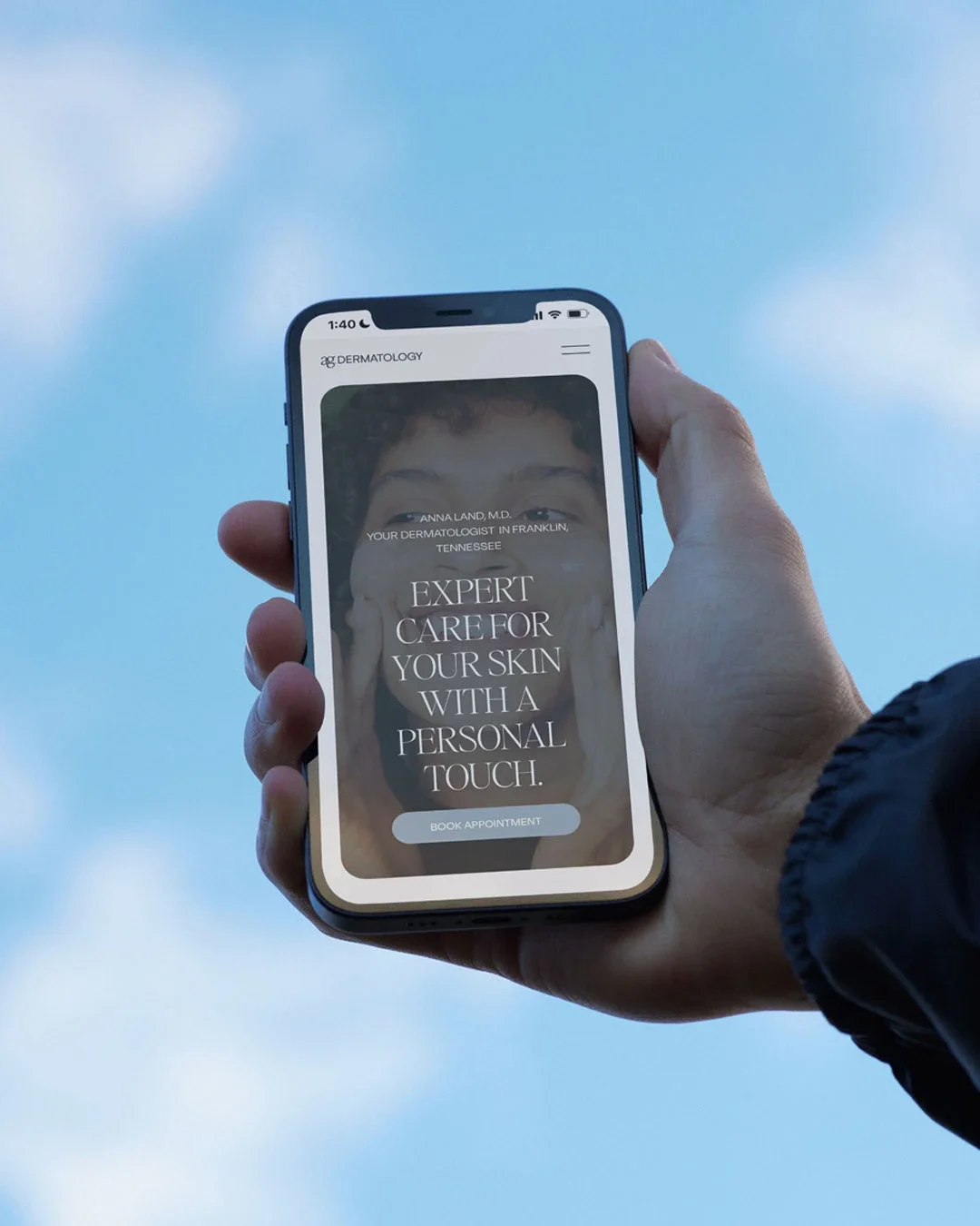 A person holding a smartphone displaying a dermatology clinic website with a smiling child on the screen. The website text reads: 'Expert care for your skin with a personal touch.'