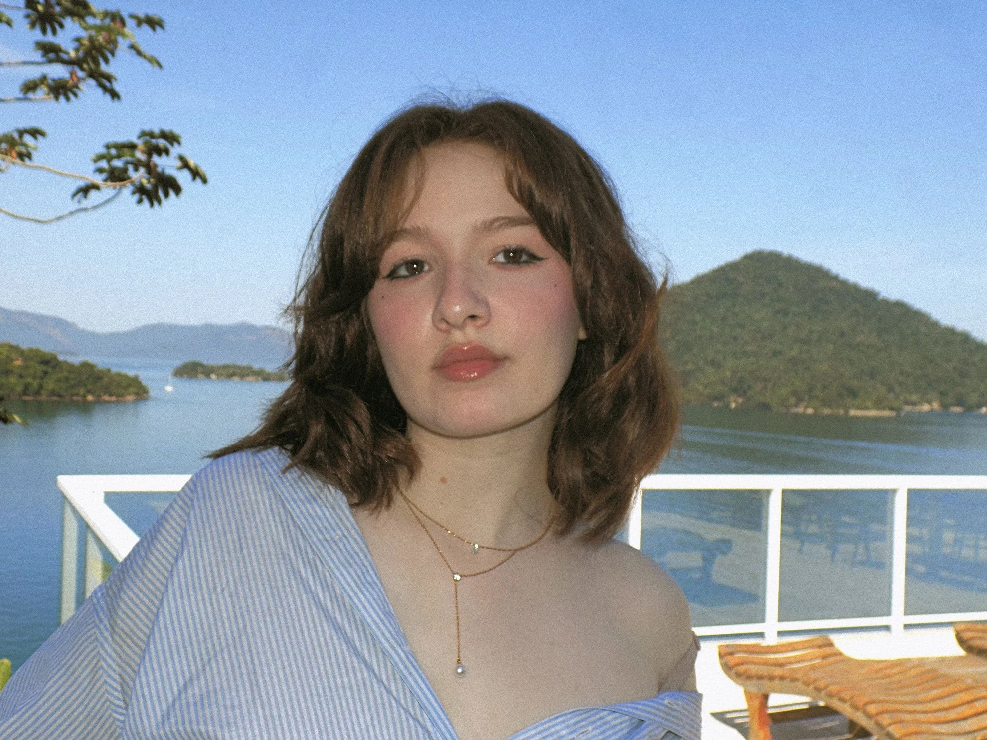 A young woman with shoulder-length brown hair, wearing layered gold necklaces and a blue and white striped shirt, posing outdoors near a body of water with mountains and greenery in the background.