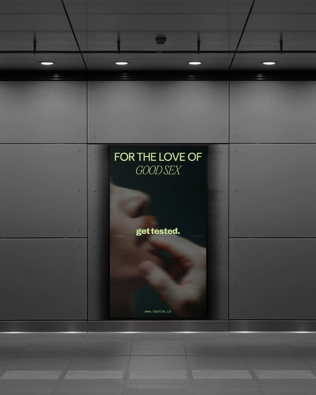 Digital billboard advertises testing for good sex with the message 'For the love of good sex, get tested' and the website www.testie.co. The billboard is inside an underground subway station with gray walls and ceiling with recessed lighting.