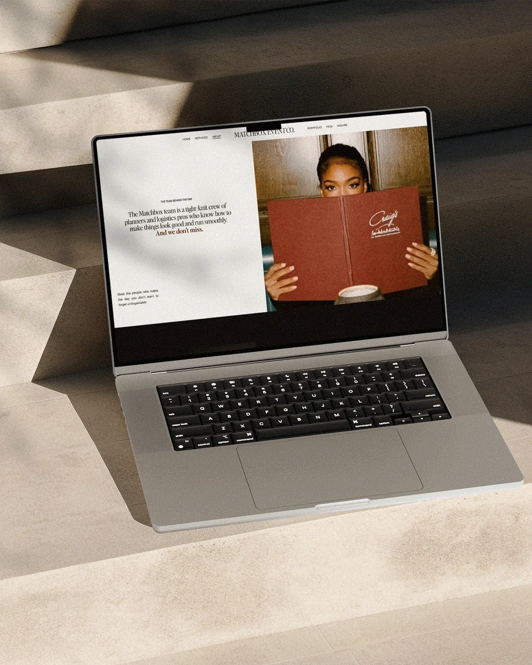 Open laptop on a beige surface displaying a website with a woman holding a red menu and a coffee cup. The website features text about the Matchbox team and a photo of a woman reading.