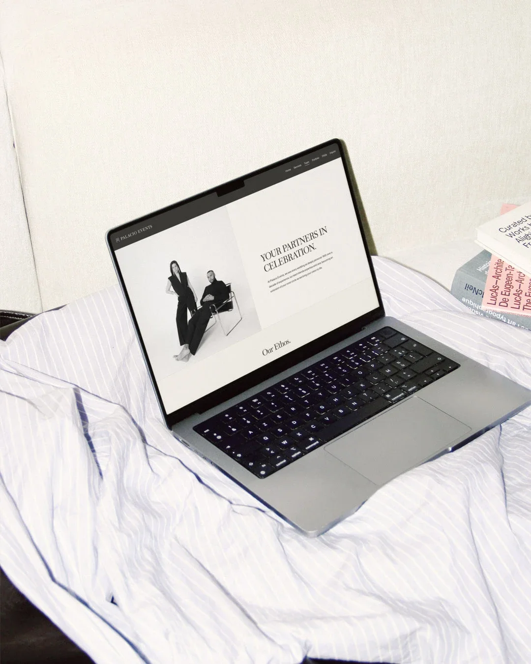 A laptop showing a website titled 'Your Partners in Celebration' with black-and-white images of two people, resting on a bed with white and blue striped sheets, and some books nearby.