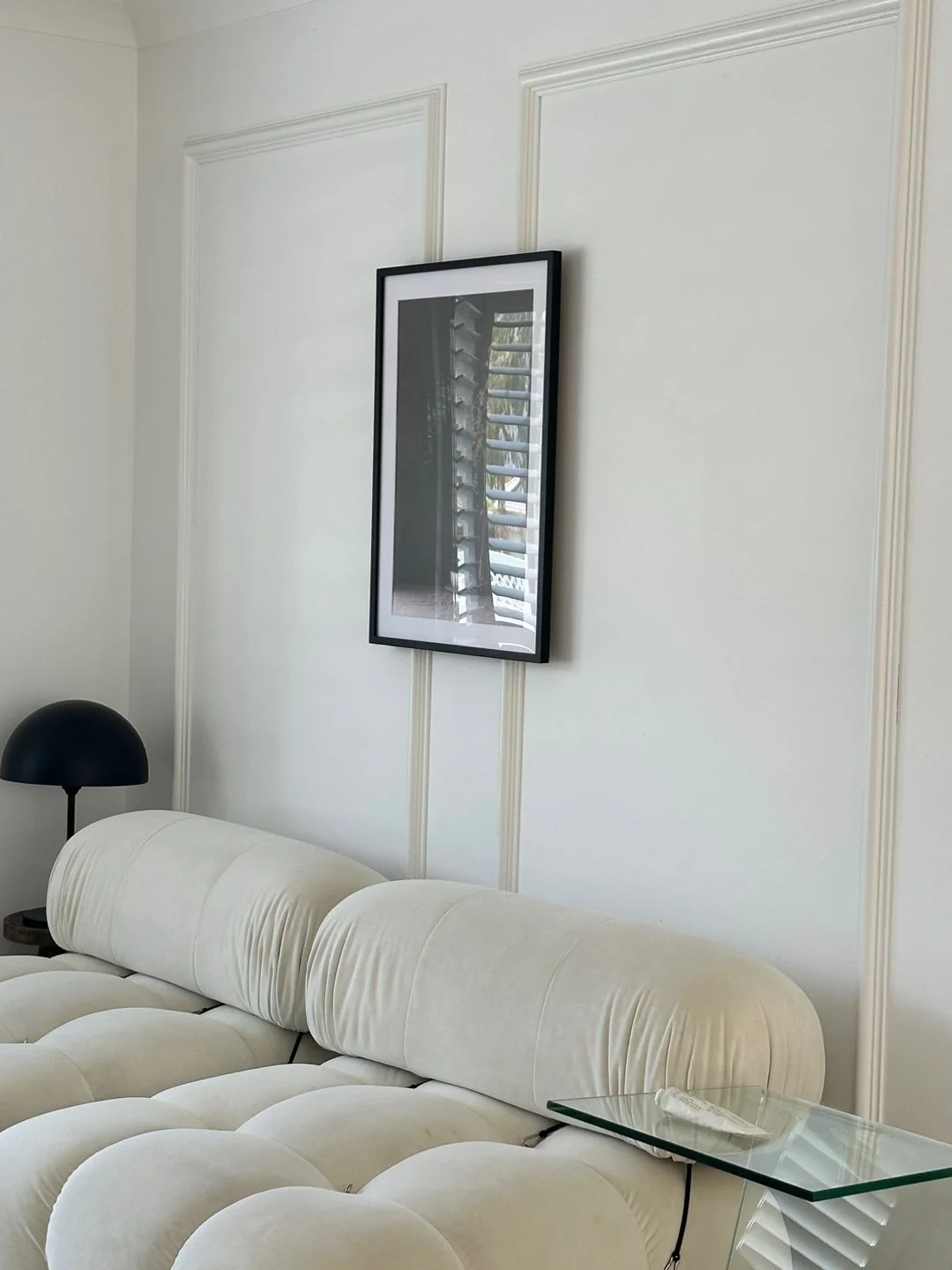 Living room with a white tufted sofa, black table lamp, framed black and white photograph of window blinds, glass side table, and white paneled wall.