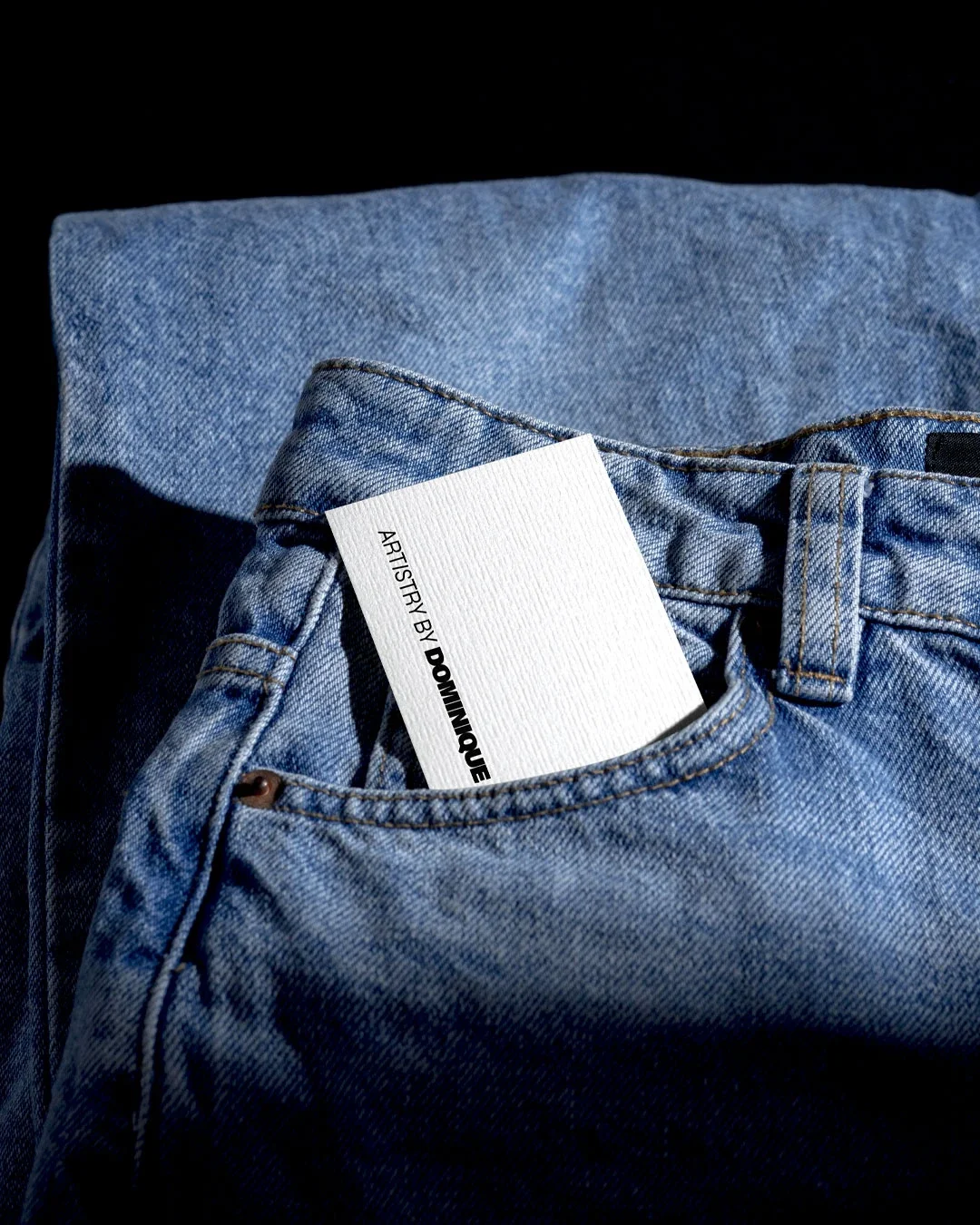 Close-up of light blue denim jeans with a white tag reading "ARTISTRY BY DOMINIQUE."