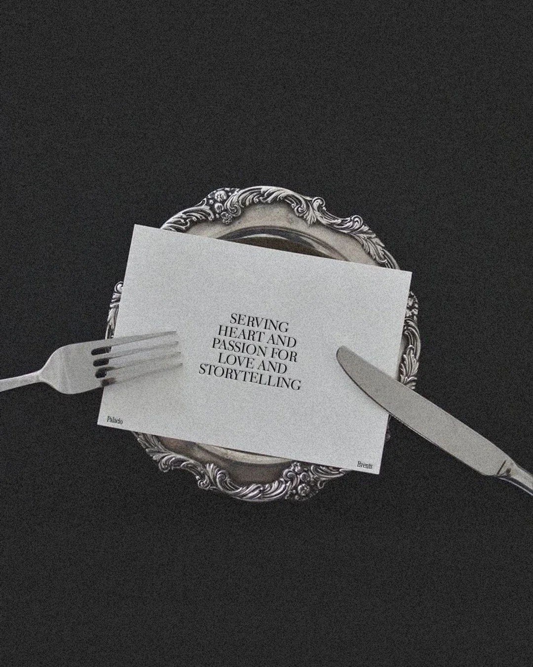 A silver ornate mirror on a black surface, with a white paper note that says 'Serving heart and passion for love and storytelling.' A fork is on the left and a knife on the right, partially obscuring the note.