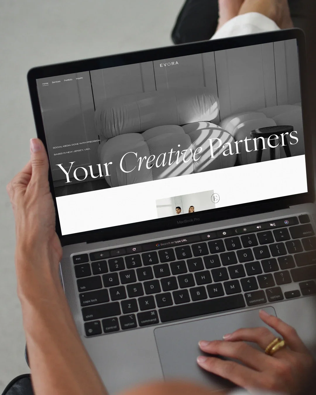 Person holding a MacBook Pro displaying a website with the text "Your Creative Partners". The website has a black and white image of a modern, minimalist interior.