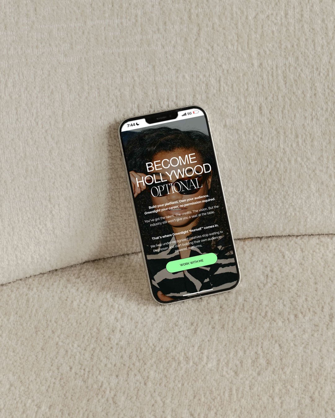 Smartphone with a promotional webpage for a platform called Greenlight, featuring the text 'Become Hollywood Optional' and a green button labeled 'Work With Me,' resting on a beige, textured fabric surface.
