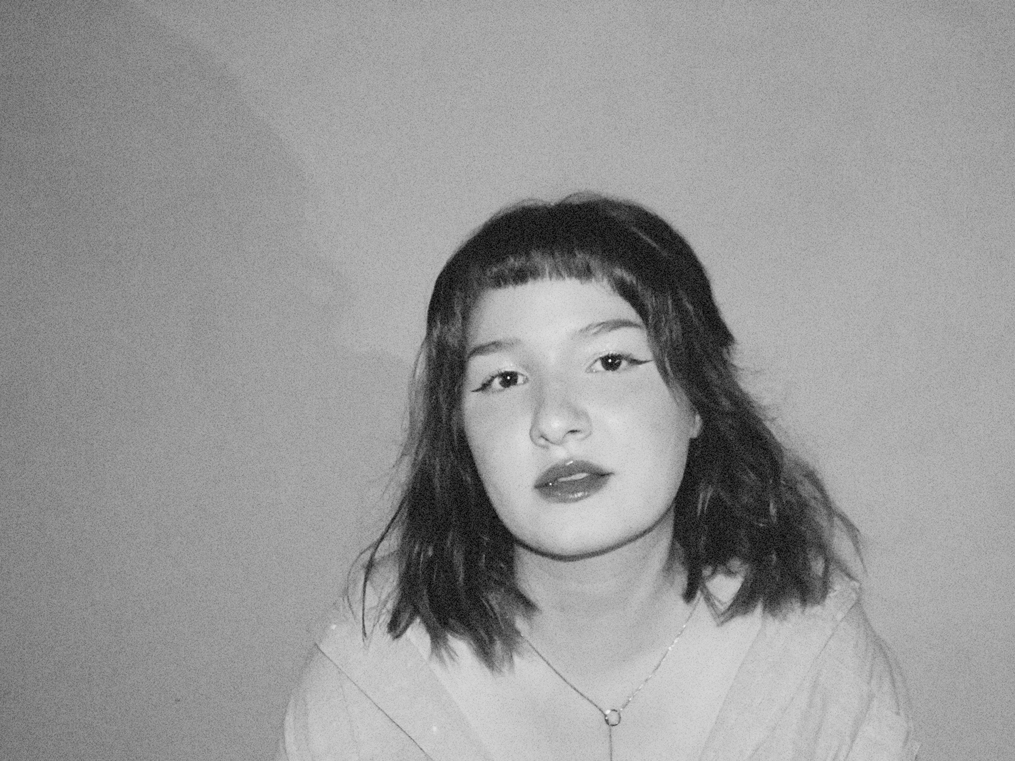 Black and white portrait of a woman with shoulder-length hair, wearing a necklace and a top with a V-neck, looking directly at the camera with a neutral expression.