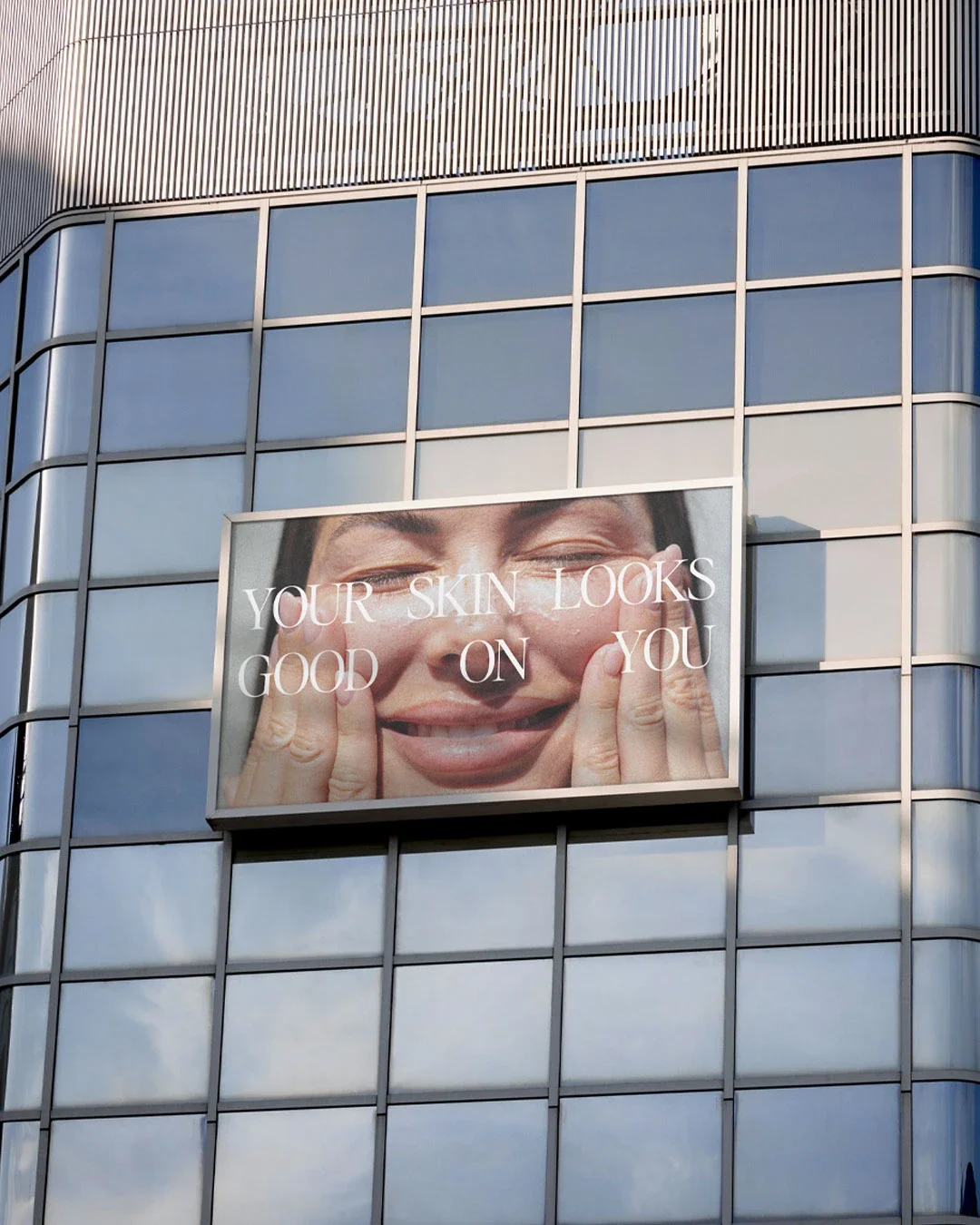 Billboard on a glass building showing a close-up of a person's face with their eyes closed and fingers on their cheeks, with the text "Your skin looks good on you" overlaid.