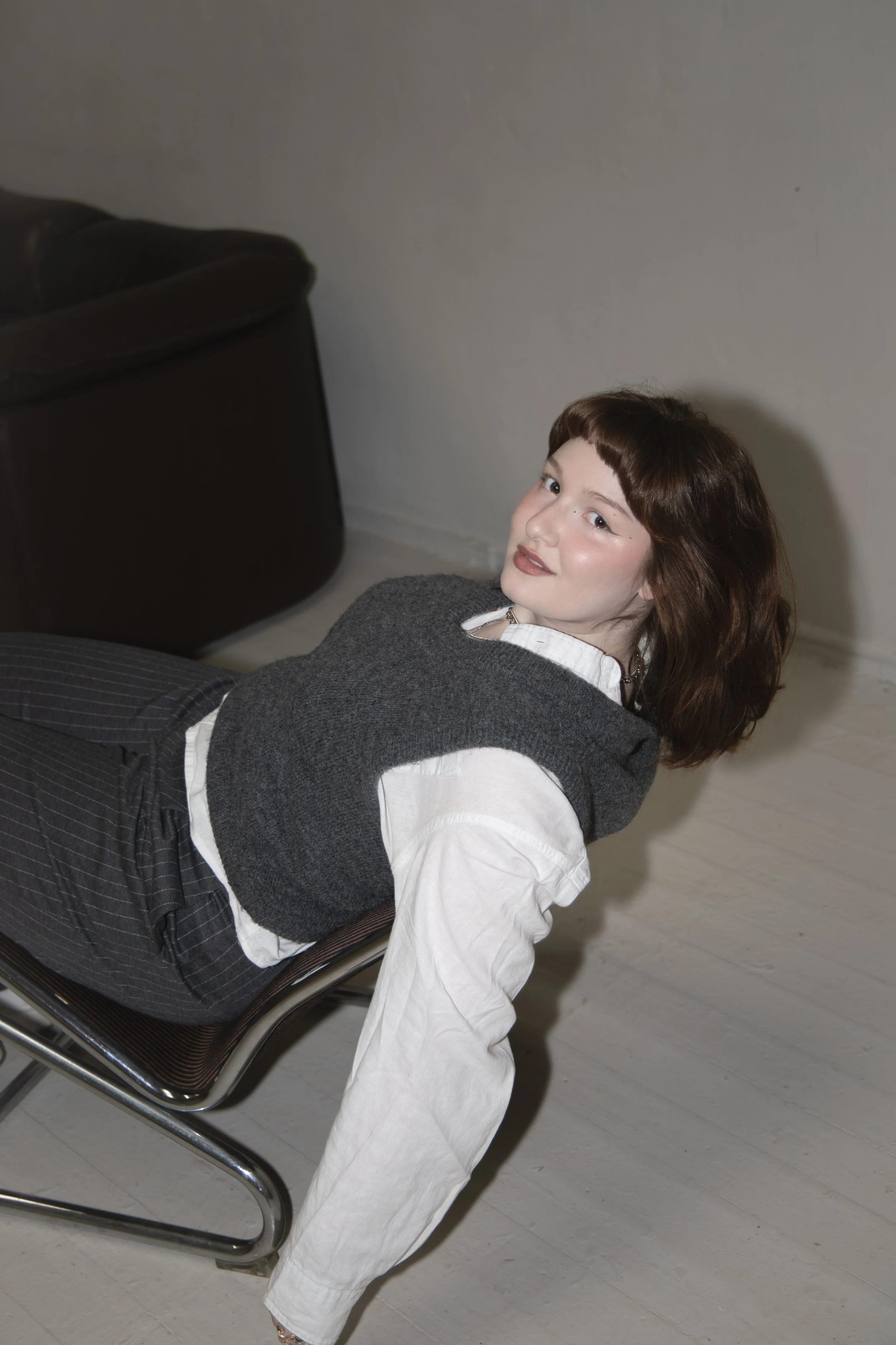 Woman with shoulder-length auburn hair, wearing a white shirt with a gray sweater vest, leaning back in a chair, looking at the camera, with a black sofa in the background.