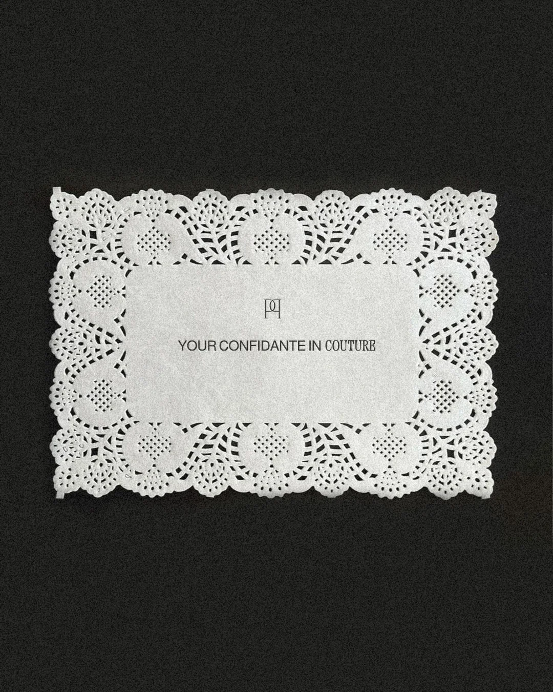 Elegant white lace doily with a rectangular message area in the center, displaying the text 'YOUR CONFIDANTE IN COUTURE' against a black background.