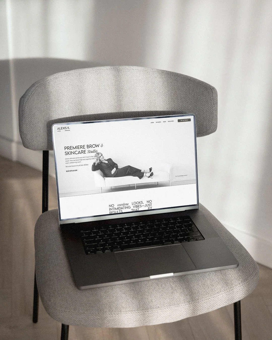 A laptop resting on a light gray upholstered chair with a black and white website displaying a woman lying on a couch on the screen.