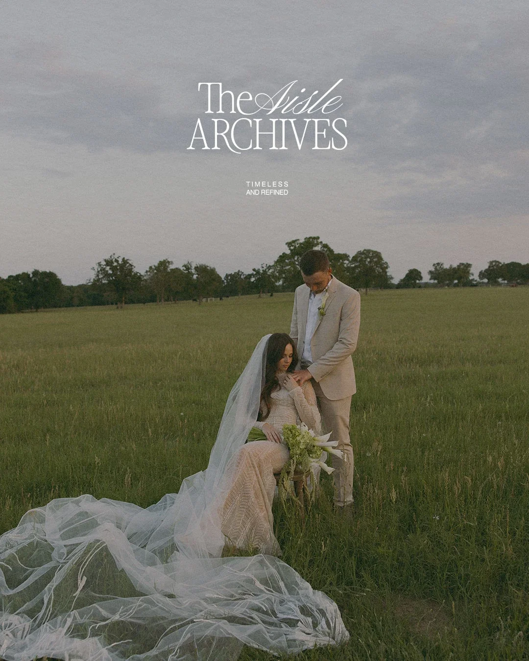 A bride and groom in wedding attire in an open field, with the groom standing and the bride sitting on a small stool, holding a bouquet of flowers, under a cloudy sky.