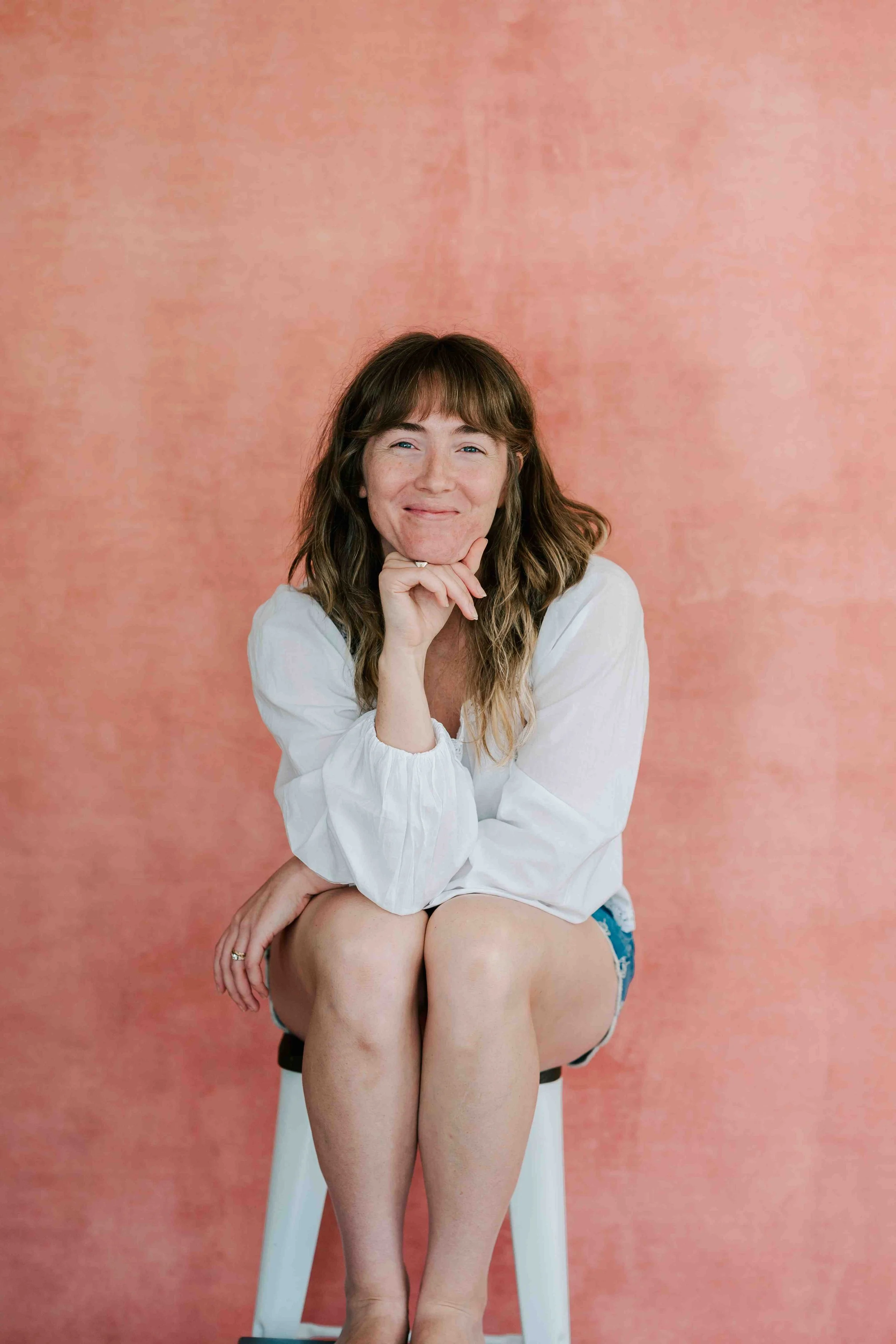 A woman sits on a chair smiling with her head resting on her hand