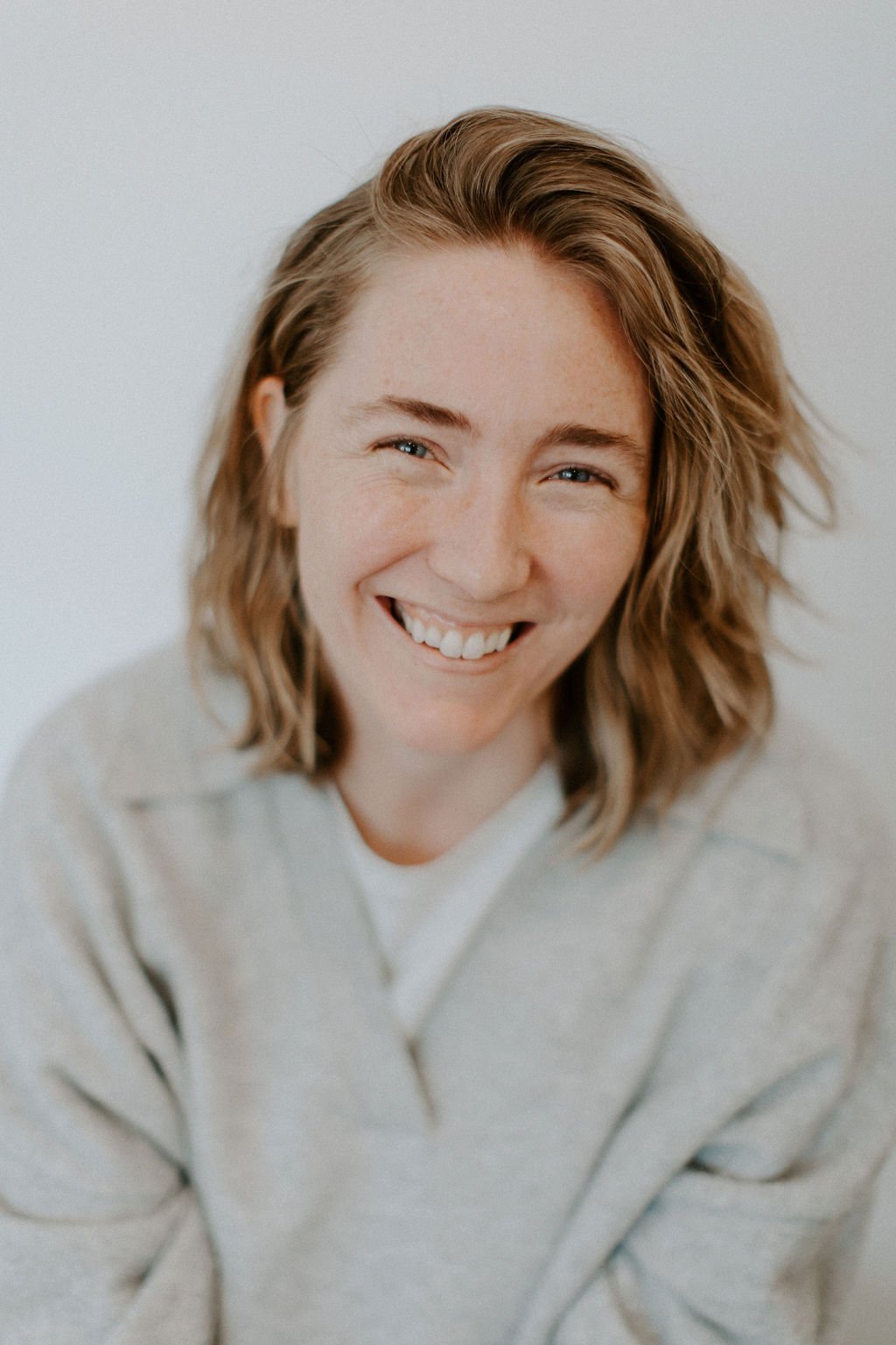 A smiling woman with shoulder-length wavy hair, wearing a light-colored sweater, looking at the camera against a plain white background.