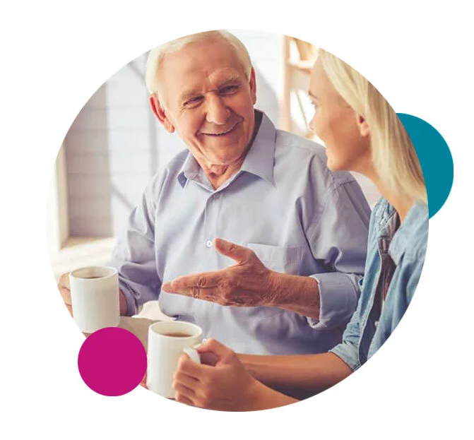 An elderly man and a woman having a conversation while sitting at a table with two coffee mugs.
