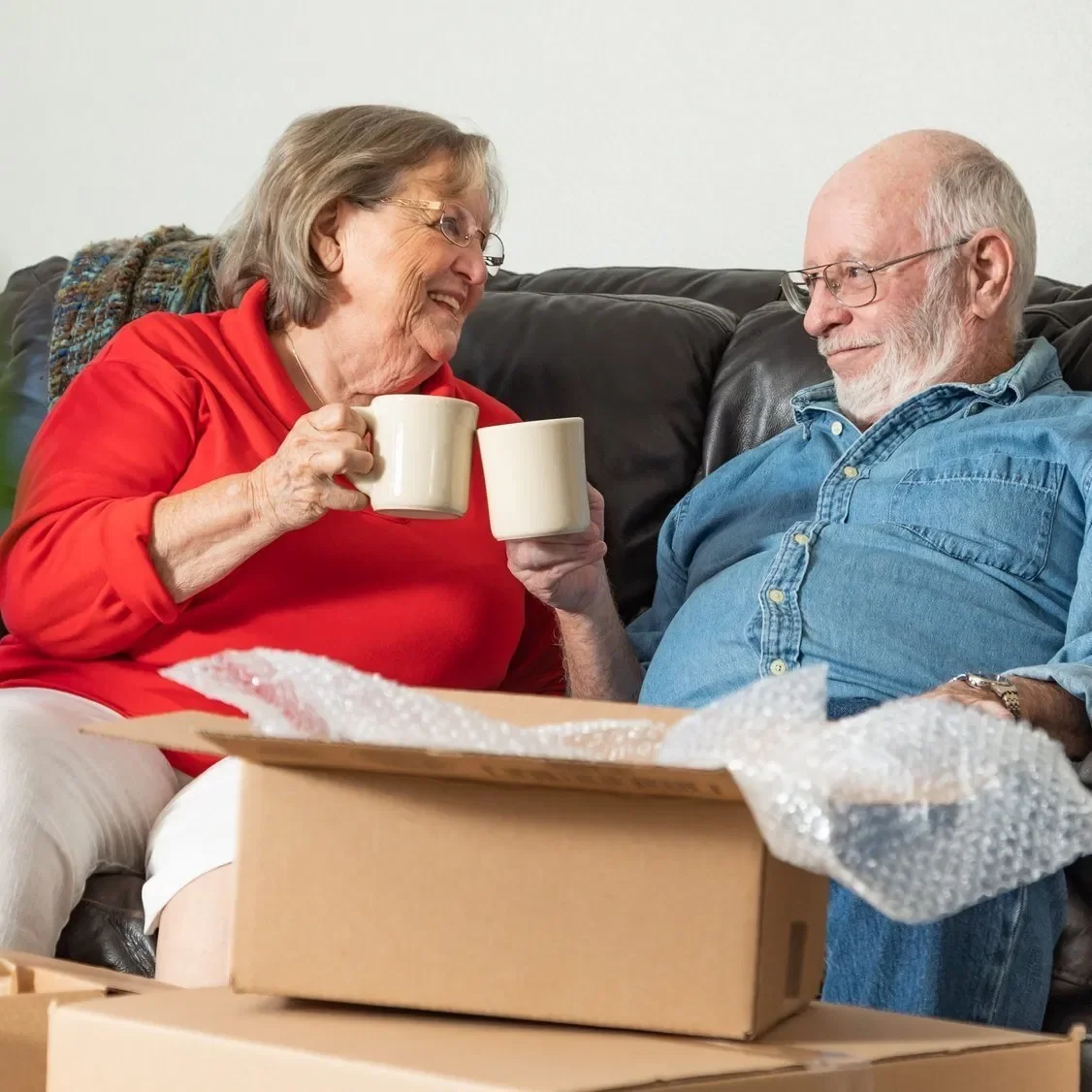 An older couple relaxes together on a sofa, sharing a cup of tea amidst moving boxes