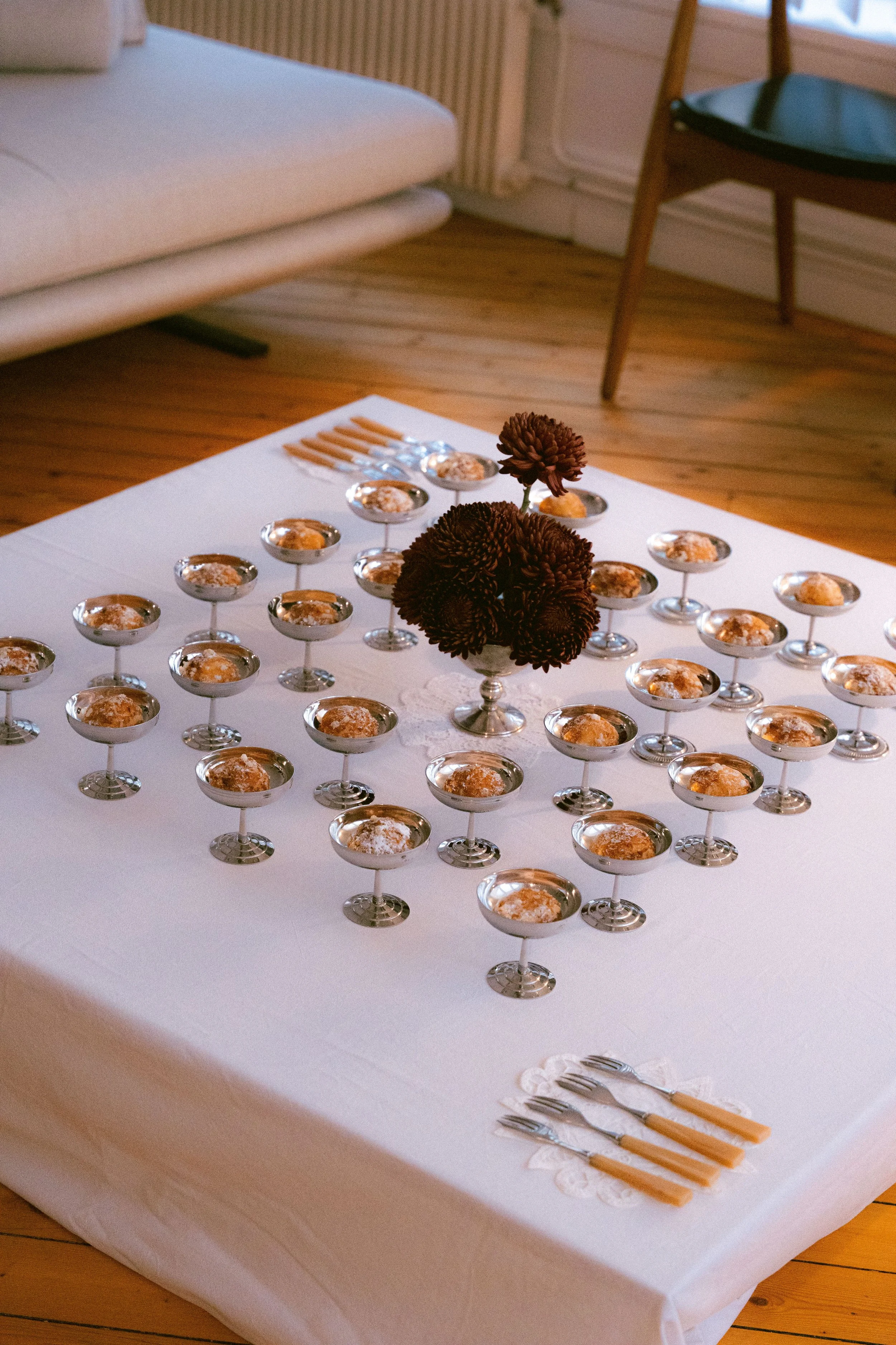 Une table buffet couverte d'une nappe blanche avec des coupes en inox, une décoration florale moderne avec des chrysanthèmes bordeaux au centre, et des couverts et napperons en dentelle ou broderie vintage.