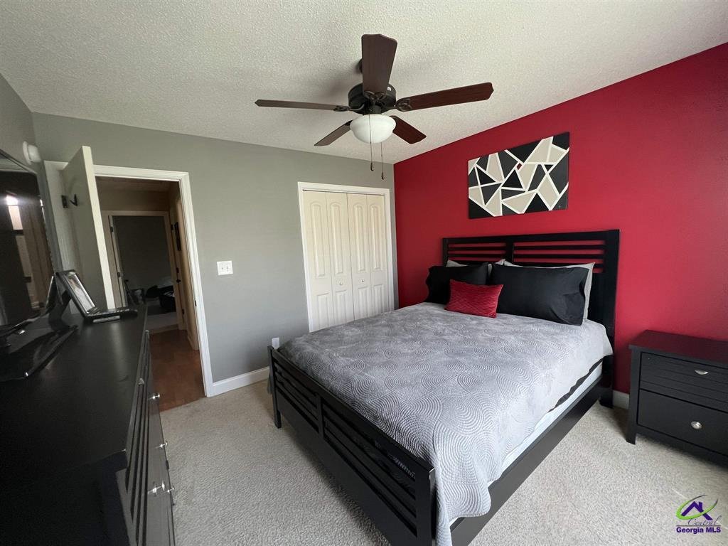 Modern bedroom with gray and red color scheme, featuring a black bed frame, gray bedding, red accent pillow, abstract wall art, ceiling fan, dresser, and nightstand.